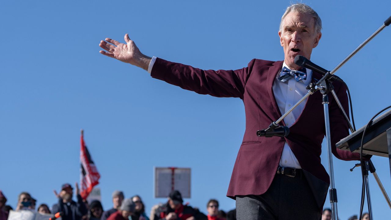 Bill Nye "The Science Guy" at a rally with his arm out stretched, speaking to the audience